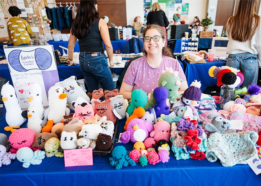 Vendor smiling behind a display table filled with colorful handcrafted crochet plushies and knitted items at a pop-up market.