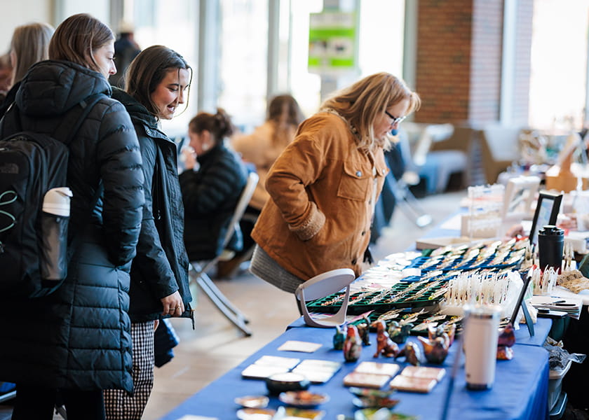 Shoppers in winter jackets bending over to examine jewelry and handmade goods on a vendor's table.
