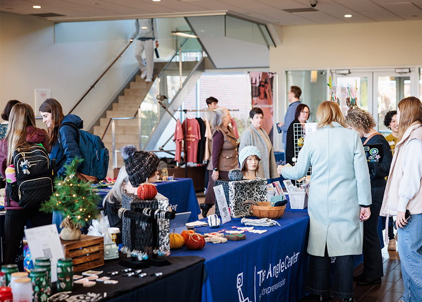 Attendees and shoppers browsing various artisan vendor tables inside a brightly lit hall at a pop-up market.
