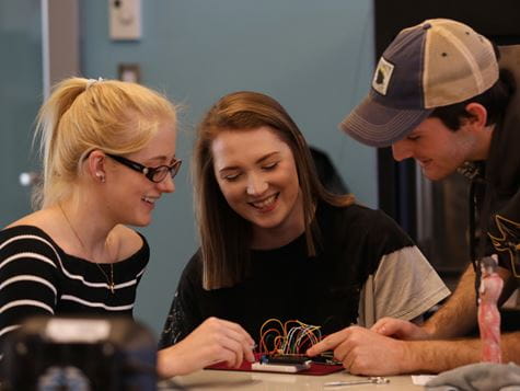 two girls and a guy looking down on a tablet on the table