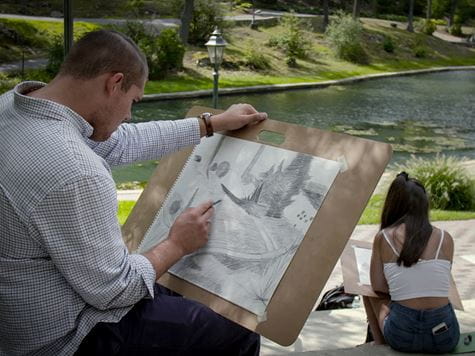 a guy sitting down outside with a board and drawing on paper