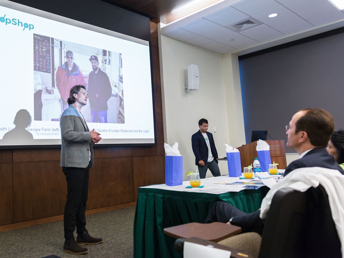 A man standing in front of a projector pitches his business idea to a panel of judges