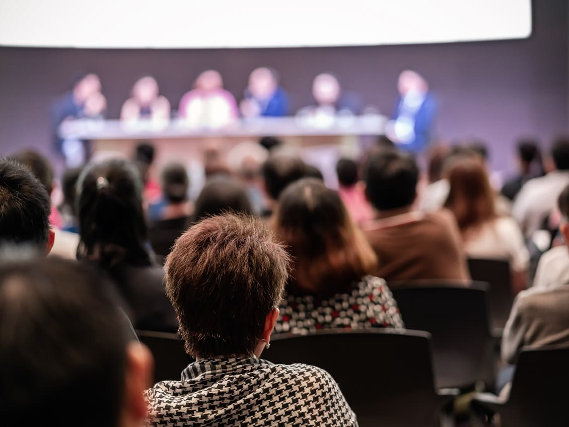 Audience members viewed from behind facing a blurred panel presentation under a bright stage screen.