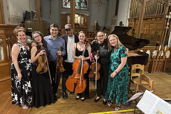 Seven musicians smiling together while holding violins and a cello near a grand piano in a church.