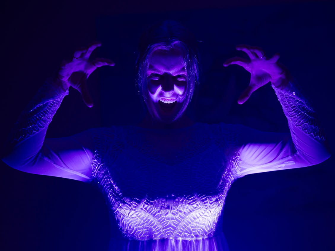 A person dressed in white raises their hands in a spooky pose, their face illuminated by eerie purple lighting against a dark background.