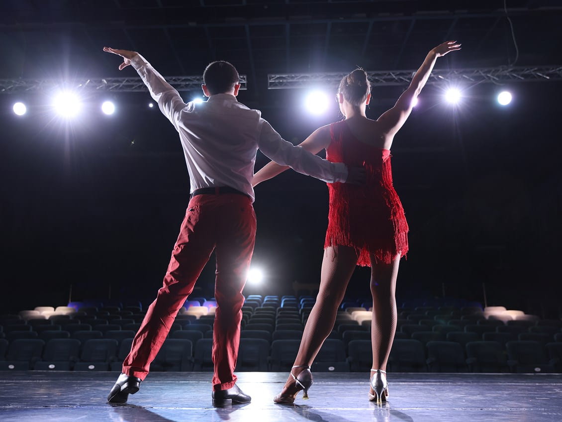 Two dancers pose onstage with their backs to the camera, one wearing a white shirt and red pants and the other a red fringe dress.