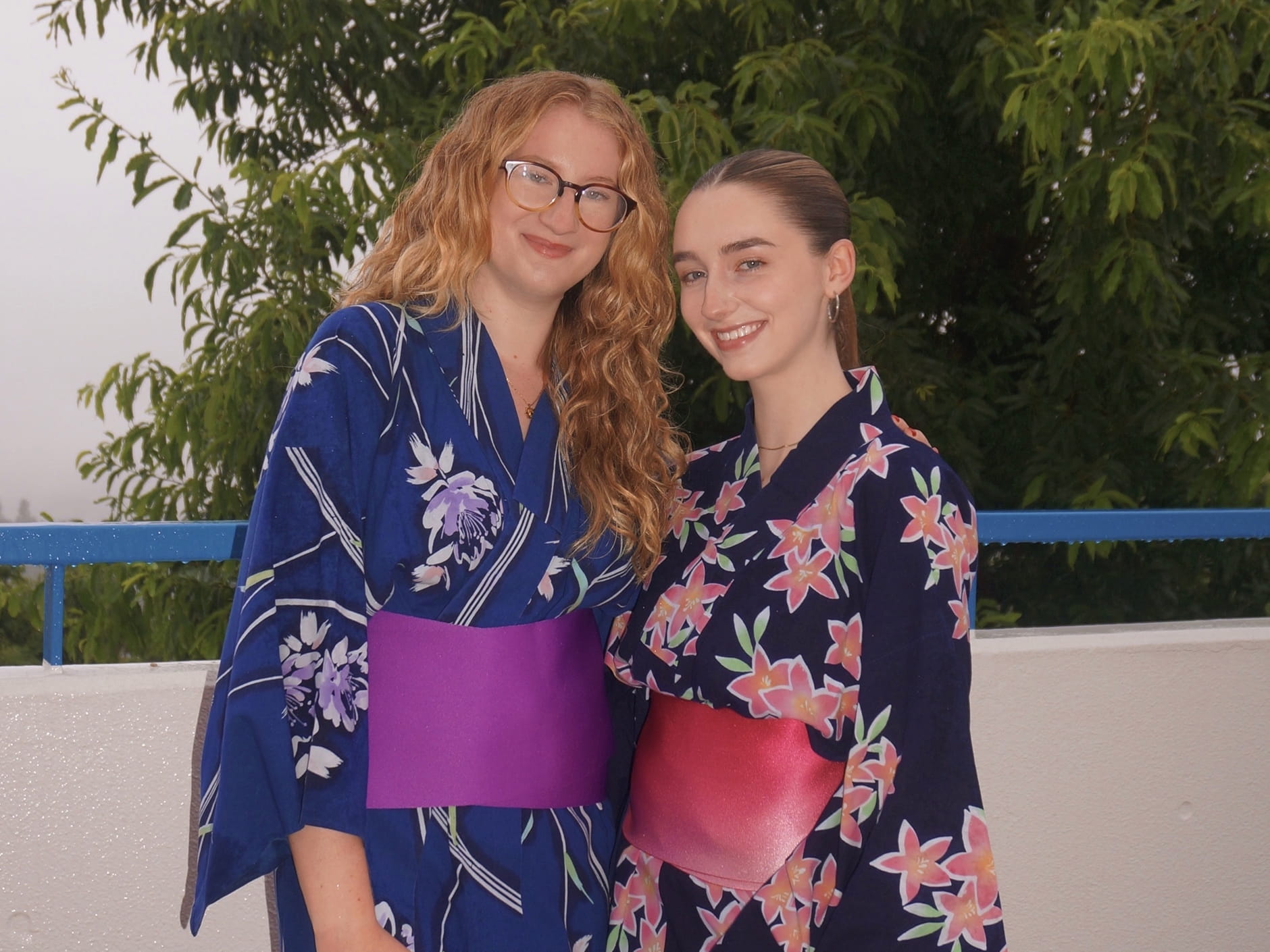 Two young women smiling in traditional Japanese kimonos with colorful obi belts against green leaves.