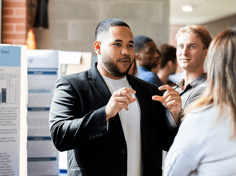 A male student in a blazer gestures while explaining his research poster to an attendee during a senior thesis presentation at Endicott College.