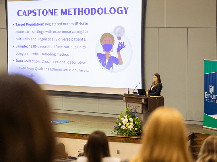 A student stands at a podium in a lecture hall at Endicott College, presenting a "Capstone Methodology" slide about a nursing study to a seated audience.