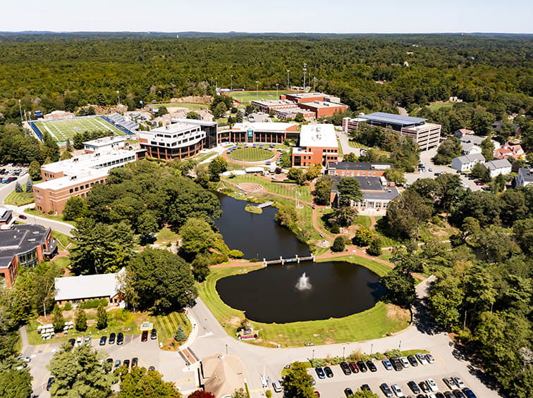 Aerial view of a wooded college campus with brick buildings, sports fields, and a central pond with a fountain.