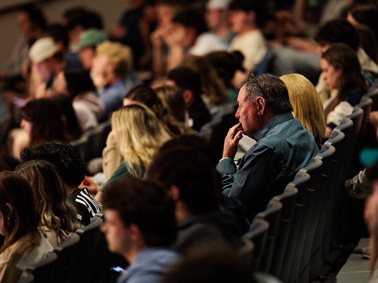 An audience sitting in a crowded auditorium, with a man in a blue shirt in profile holding his hand to his chin intently watching a presentation.