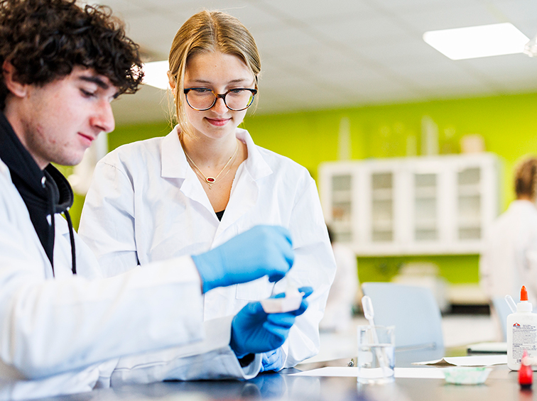Two lab students wearing coats and blue gloves working together on a chemistry experiment.