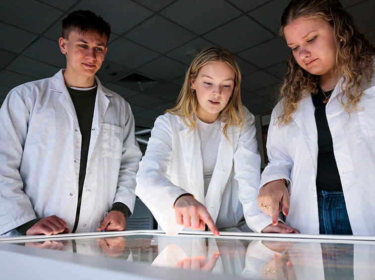 Three students in white lab coats gathered around an interactive touch table in a science lab.