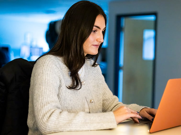 A young woman with long dark hair wearing a white cardigan sits at a desk working on a laptop.