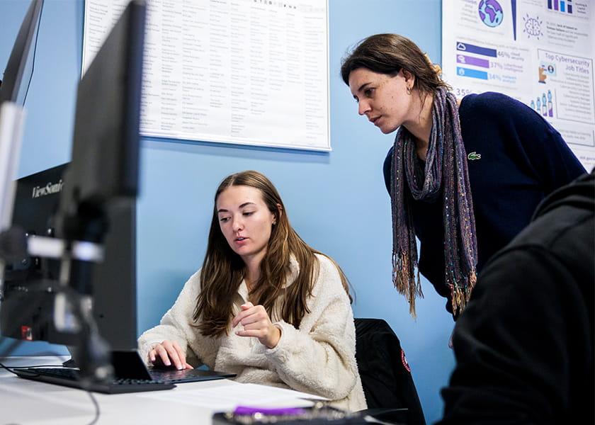 A woman wearing a colorful scarf leans over to assist a student sitting at a computer in a classroom.