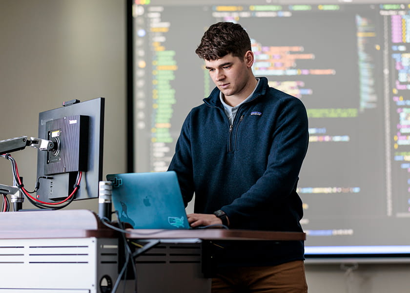 A young man standing at a podium working on a laptop in front of a large screen displaying software code.