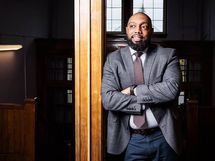 A smiling man with a beard wearing a grey plaid suit jacket, tie, and blue trousers, leaning against a wooden doorframe with arms crossed.
