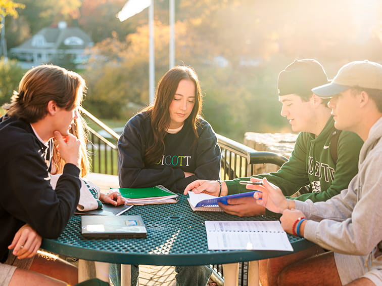 Four Endicott College students sitting around an outdoor patio table studying together in the sunlight.