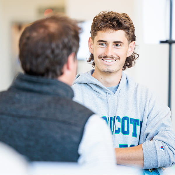 A young man wearing a grey hooded sweatshirt smiling while talking to another person whose back is facing the camera.