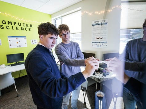 Two students in a computer science lab looking at a glass panel while interacting with virtual reality controllers.