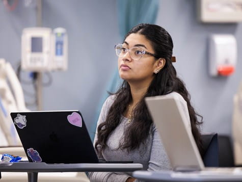A female student with glasses and long dark hair sitting thoughtfully with her laptop in a healthcare lab setting.