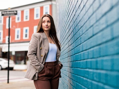 A woman dressed in a plaid blazer standing outside, leaning against a bright blue brick wall.