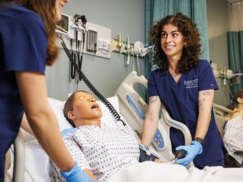 A female nursing student with curly hair smiles while taking blood pressure on a medical training mannequin.