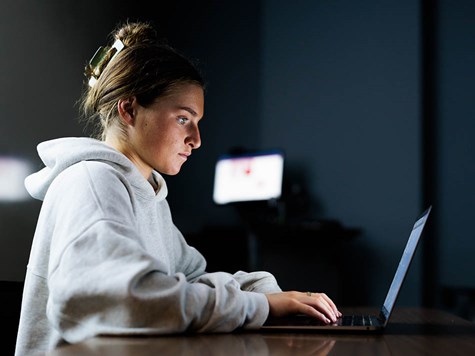 A female student in a gray hoodie sitting at a table and focused intently on her laptop.