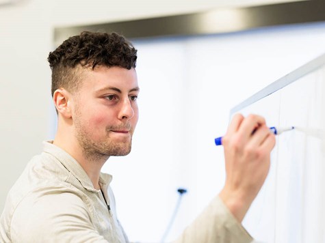 A male student with short curly hair writing on a whiteboard with a blue marker.