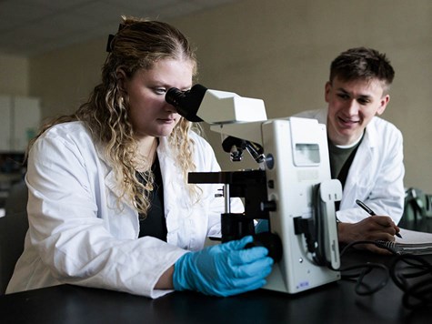 A female student in a lab coat using a microscope while a male classmate takes notes beside her.