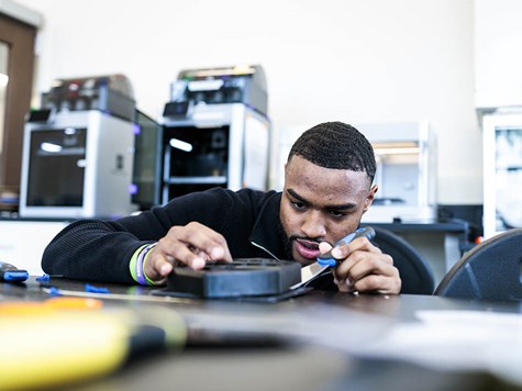 A male student carefully using a scraper tool on a 3D-printed part in an engineering lab.