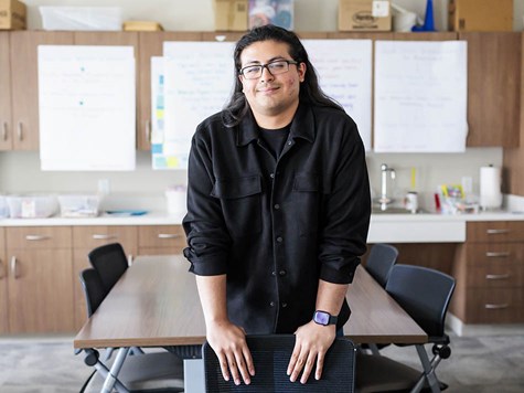 A male teacher with long dark hair leaning against a chair in a classroom and smiling.