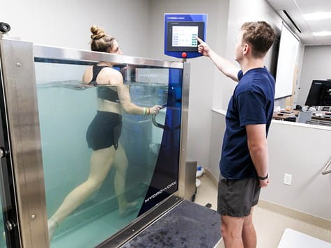 A woman walking on an underwater treadmill inside a glass tank as a man operates the control panel.
