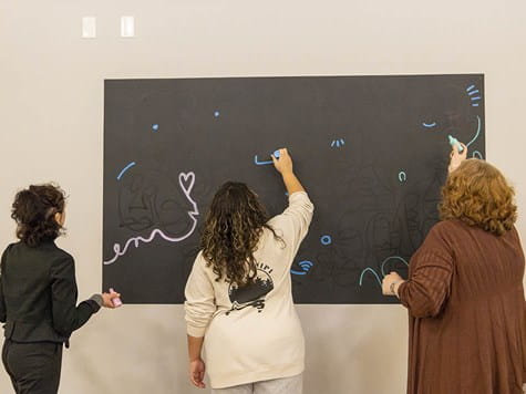 Three women standing side-by-side drawing with chalk on a large black wall in a studio.