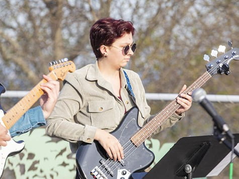 A musician wearing sunglasses and a corduroy jacket playing an electric bass guitar outdoors.